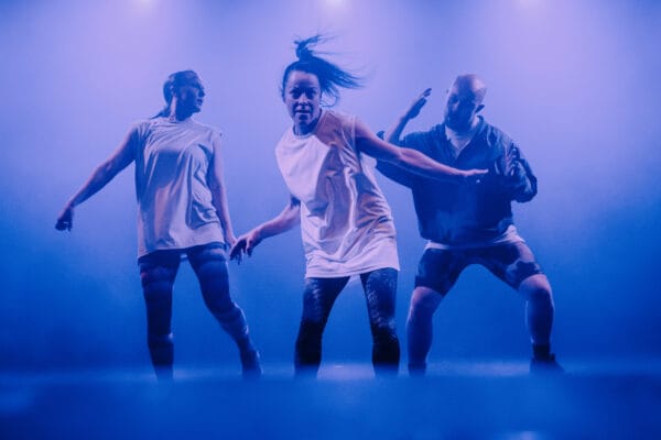 Kolme tanssijaa tanssii sinertävässä valossa.